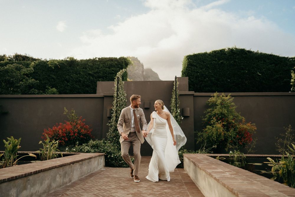 A couple walks up outdoor garden steps in wedding attire against a stone wall.