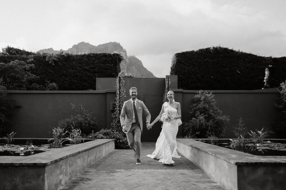 Two people walk hand in hand along a garden pathway with mountain peaks and formal hedges in the background.