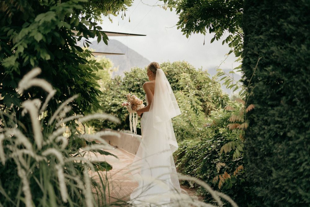 A bride in a flowing white dress and veil stands on a garden path surrounded by lush greenery.