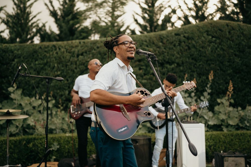 Musicians perform outdoors with acoustic guitar in front of manicured hedges.