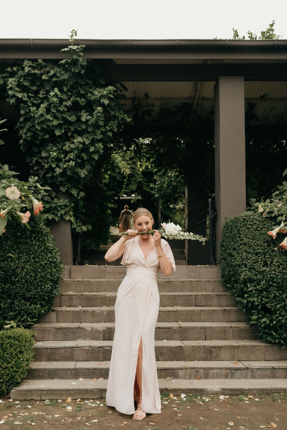 A person in an elegant white wedding dress poses on stone steps surrounded by manicured hedges and greenery.