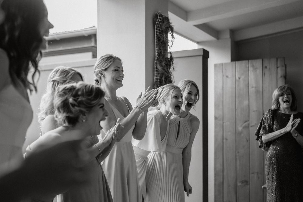 A black and white candid moment shows a group reacting joyfully during wedding preparations.