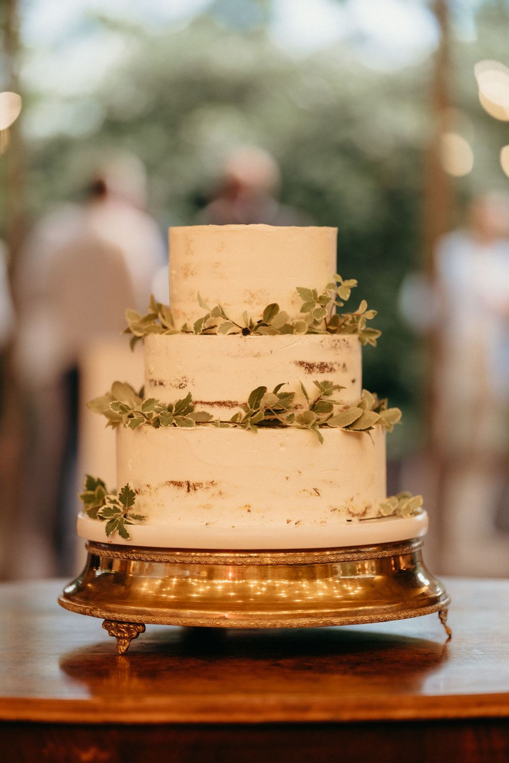 A rustic three-tiered white wedding cake with green foliage decorations sits on an ornate copper-colored cake stand.
