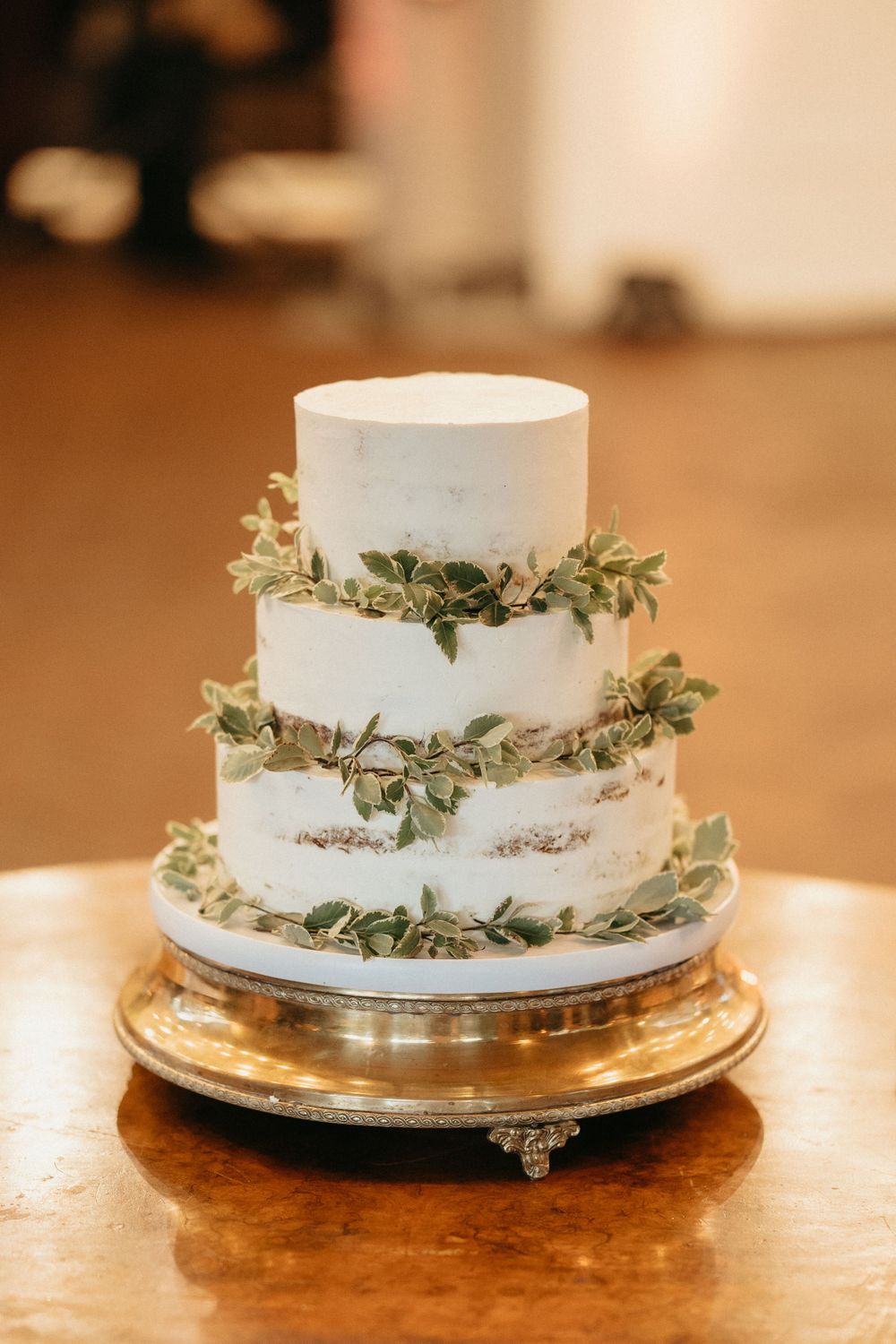 Three-tier white wedding cake decorated with delicate greenery garlands on golden stand.