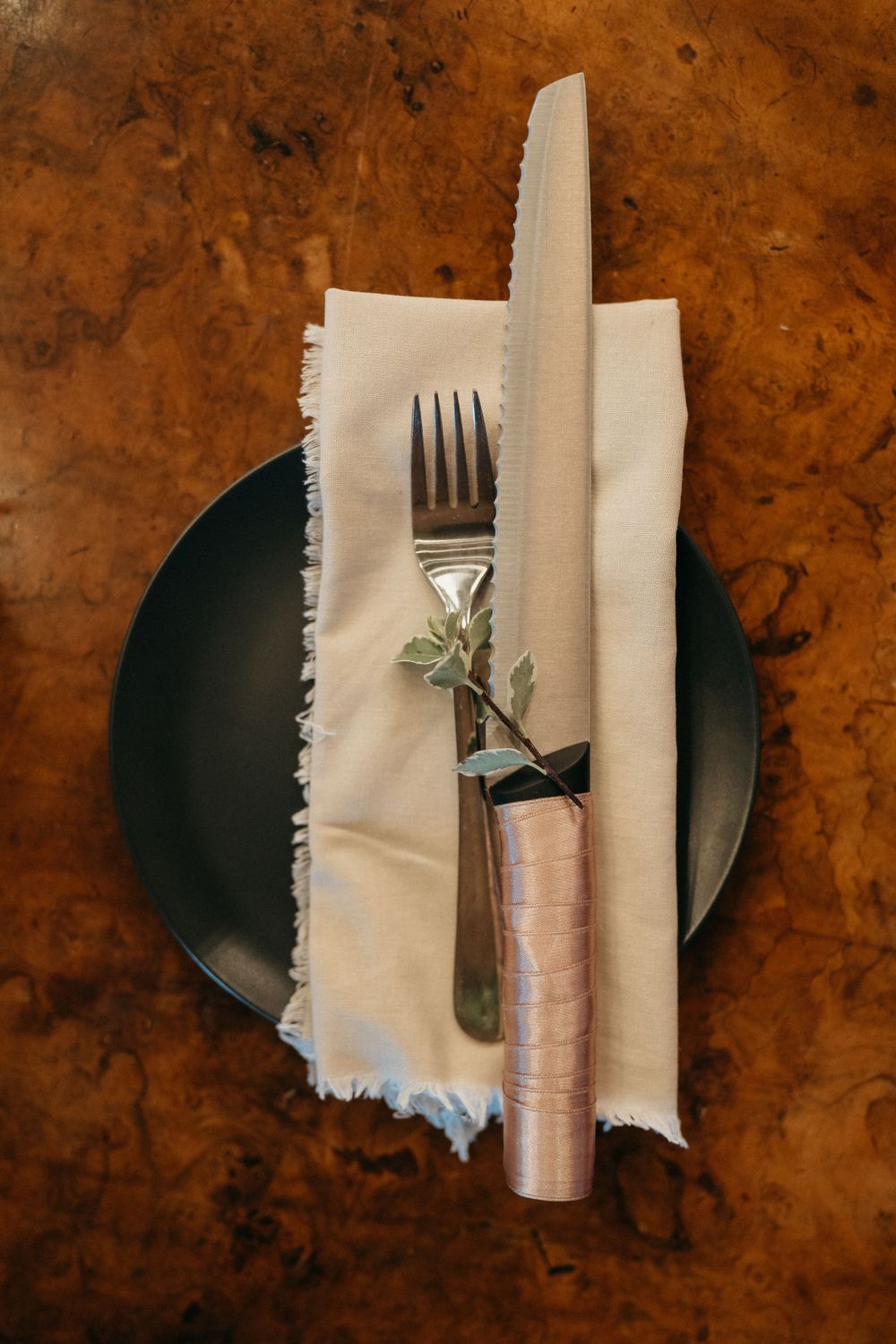 Elegant place setting with silverware wrapped in fabric on a dark plate against rustic wood surface.