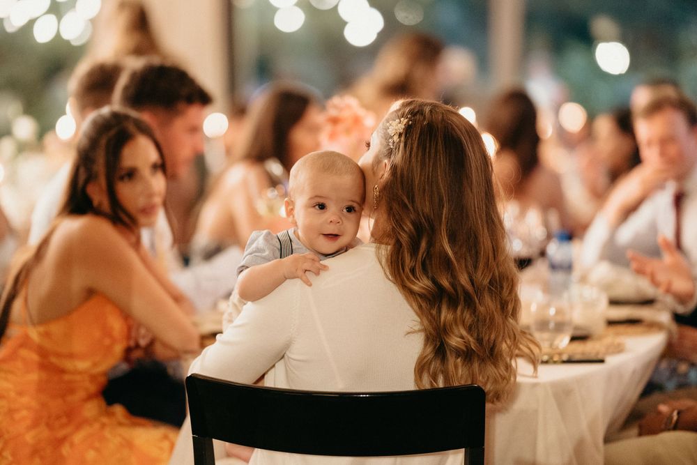 A baby peeks over a parent's shoulder during a wedding reception dinner with guests seated at tables.