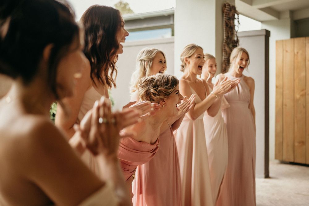 Bridesmaids in pink dresses gather excitedly in a bright indoor space before a wedding ceremony.