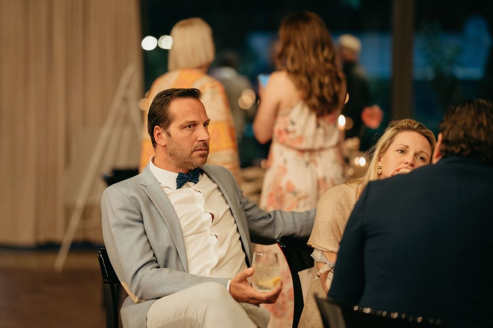 Wedding guest in light gray suit and bowtie sits at reception table.