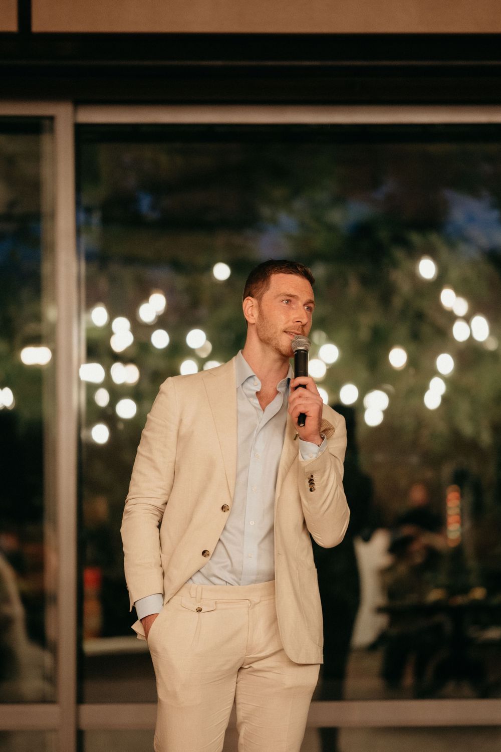 Wedding speaker in cream suit illuminated by twinkle lights during evening reception.