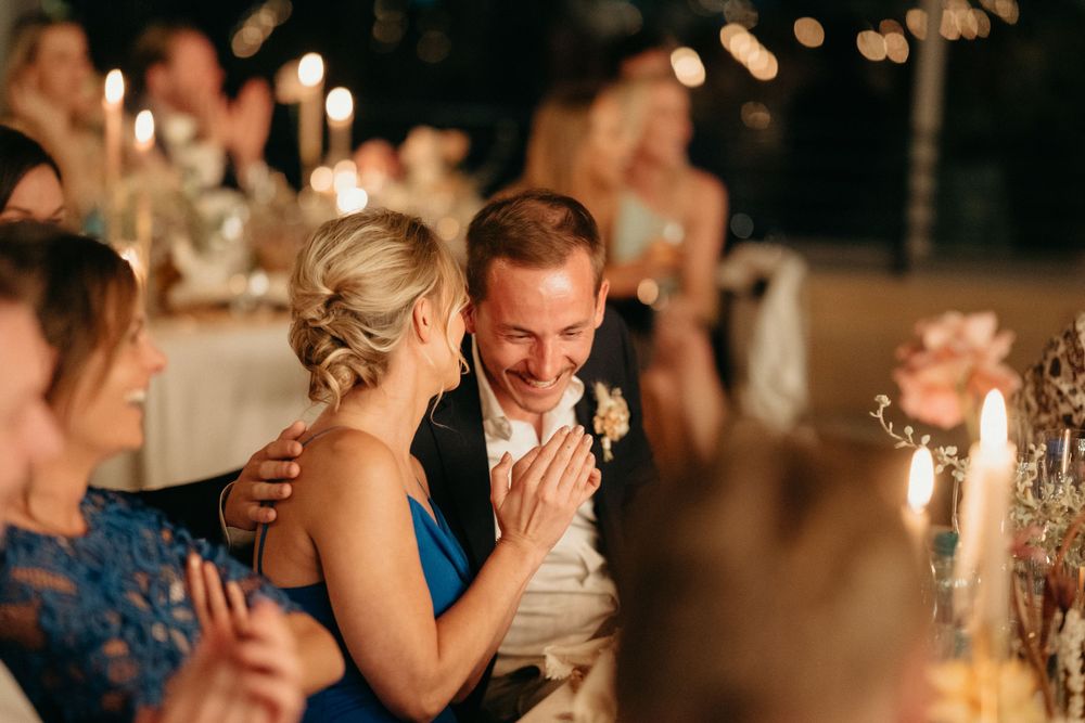 Warmly lit candlelit reception scene shows guests sharing joyful laughter at elegant wedding dinner.