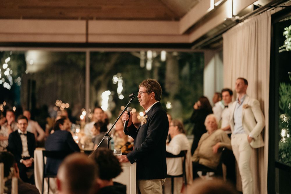 A speaker addresses wedding guests during an evening reception with large windows and foliage in background.