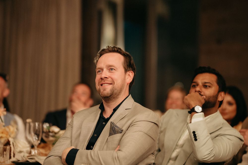 Wedding guests react with joy and laughter during reception speeches in a dimly lit venue.