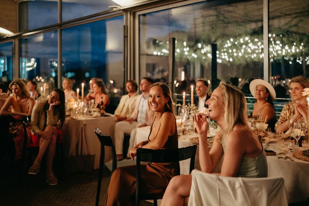 Wedding guests seated at elegant tables during reception dinner in modern venue with glass walls.