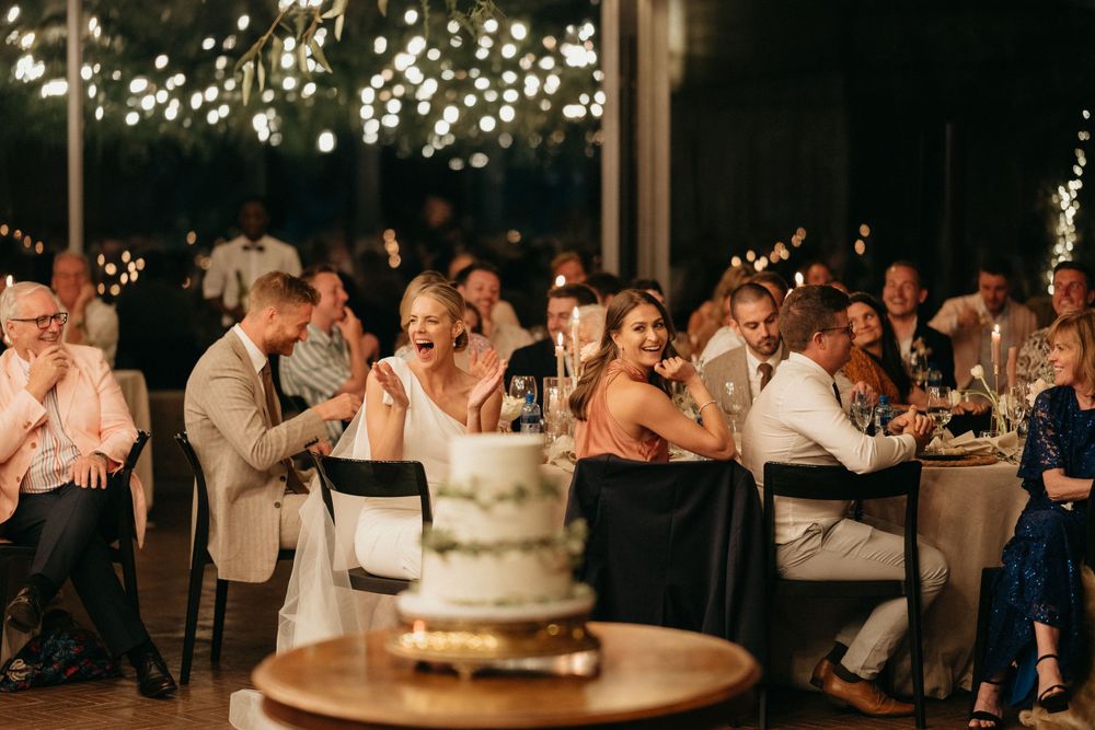 Wedding guests celebrate at reception venue with twinkling lights and wedding cake displayed on rustic wooden table.