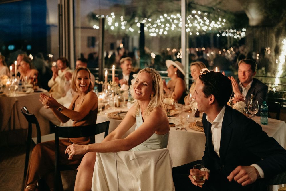 Wedding reception guests laughing and celebrating at tables decorated with string lights.