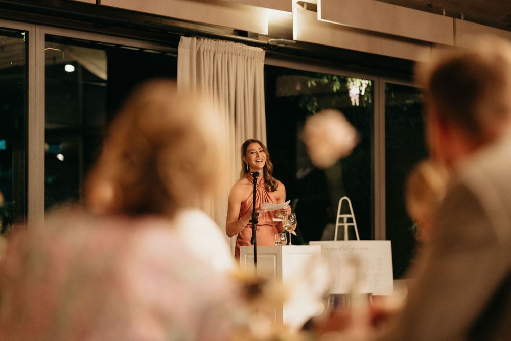 A speaker stands at a podium delivering remarks during an indoor event with soft lighting and curtains in the background.