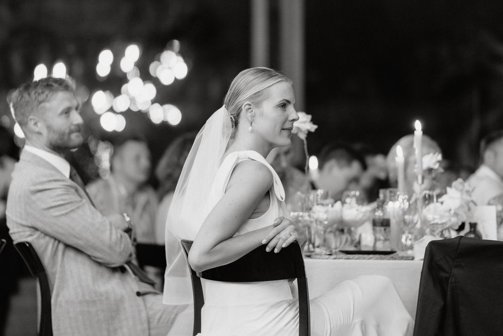 Black and white candid moment captured during evening wedding reception with bokeh lights in background.