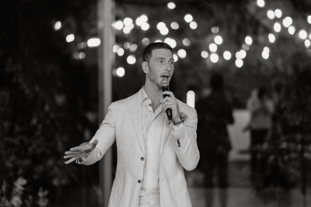 A performer in a white shirt speaks into a microphone against a bokeh light background at an evening event.