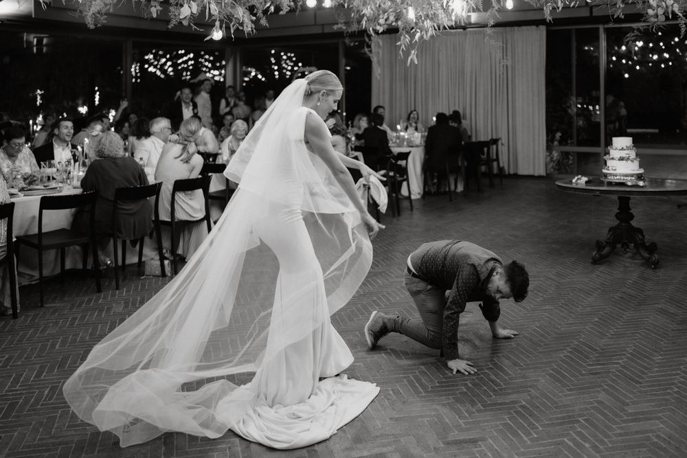 A bride in a flowing white gown and veil shares a playful moment during a wedding reception.