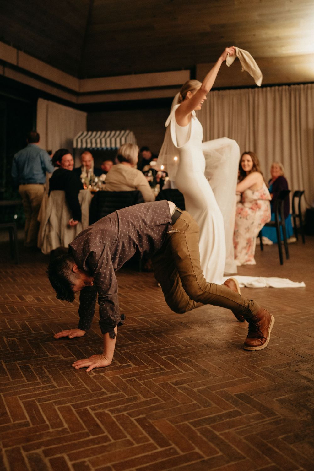 Wedding guests break dancing on a parquet floor while others watch from the sidelines at a celebration.