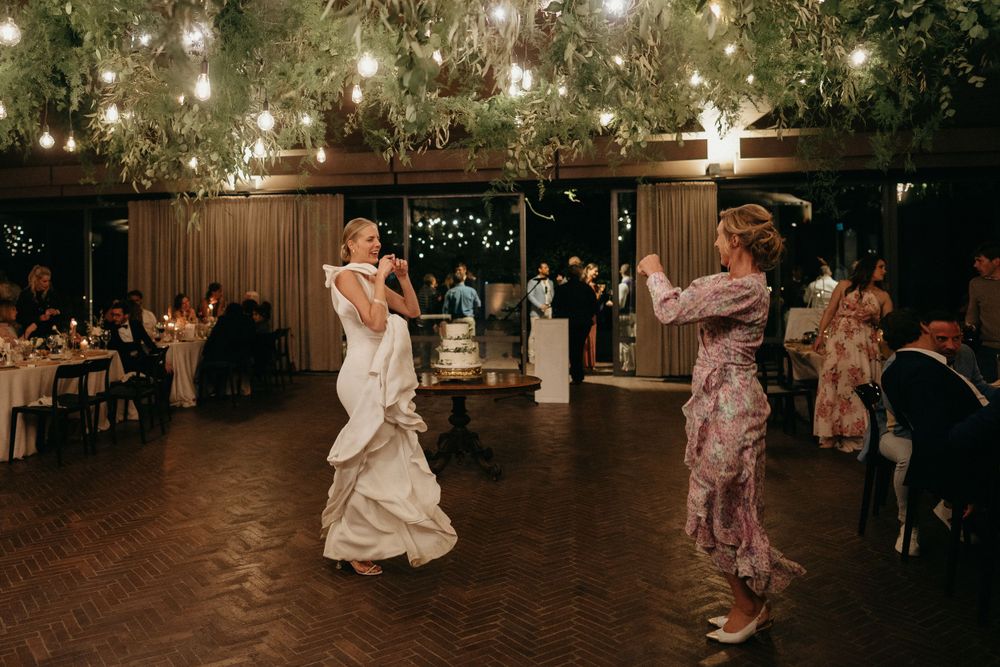 Wedding guests dance together in a rustic reception venue with twinkling lights overhead and wooden walls.