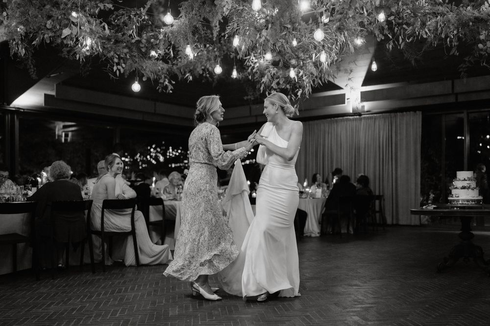 Two people dance together on an illuminated reception floor beneath hanging greenery.