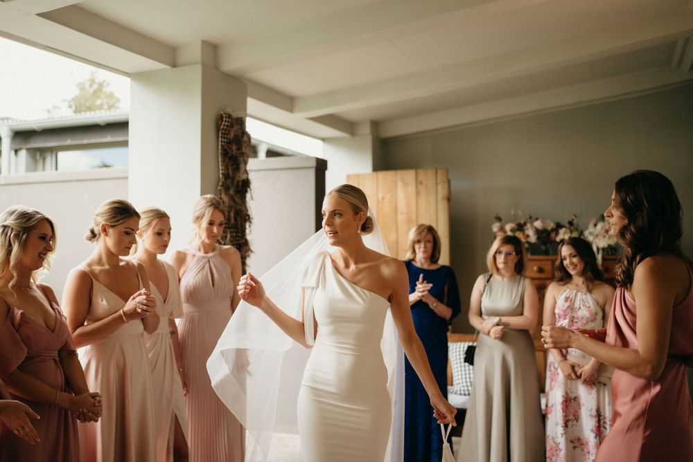 A group wearing pastel bridesmaid dresses gather around a bride in an elegant white gown.