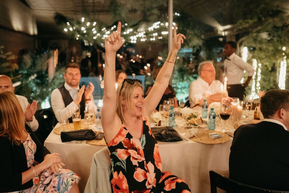 Wedding guests celebrating enthusiastically at a reception with string lights and elegant table settings.