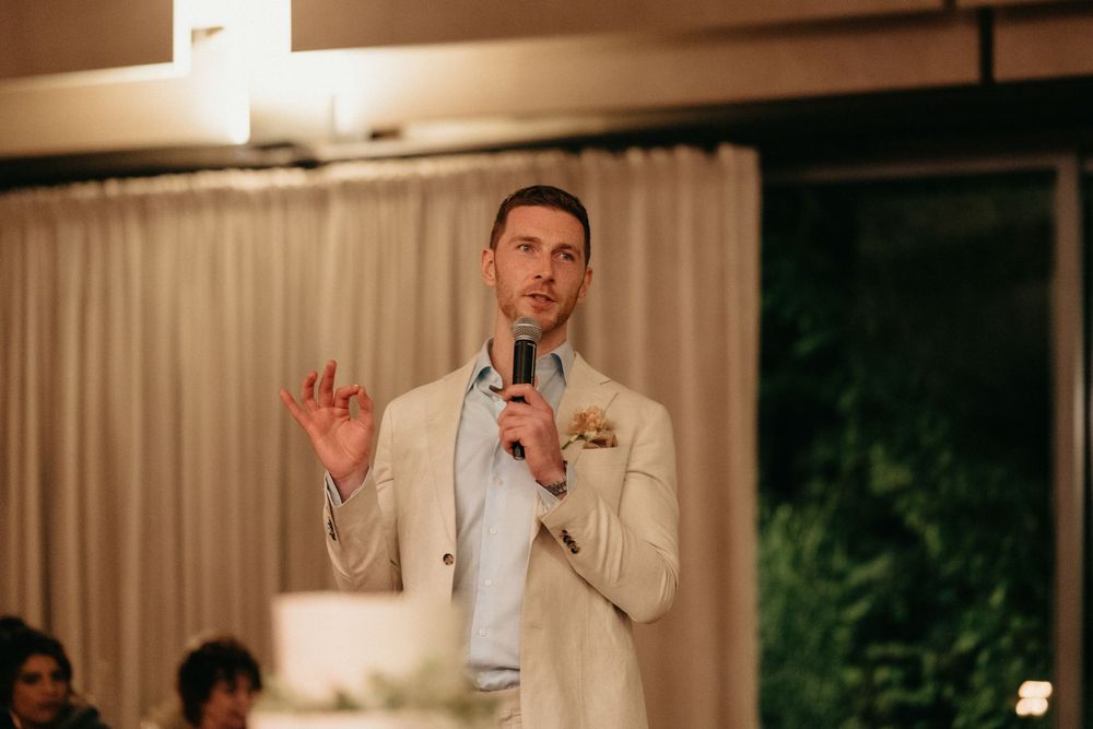 Speaker in light-colored suit giving a speech at an elegant indoor wedding venue.