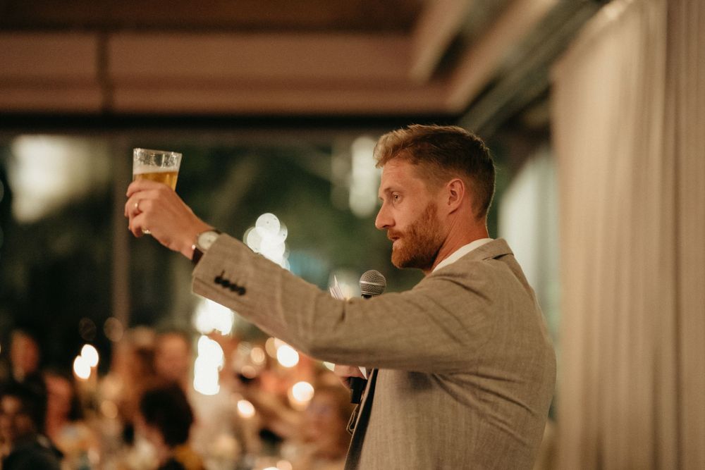 Person in grey suit raises a glass for a wedding toast at an evening reception.