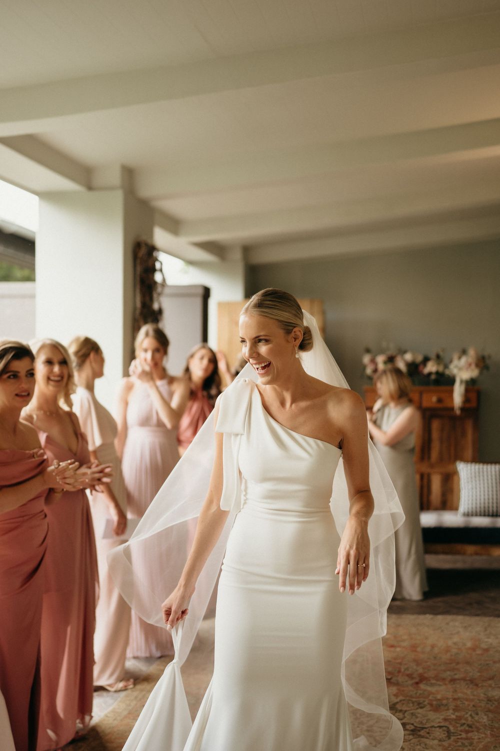 A bride in an elegant one-shoulder white wedding gown and veil twirls while her bridesmaids in pink dresses look on joyfully.