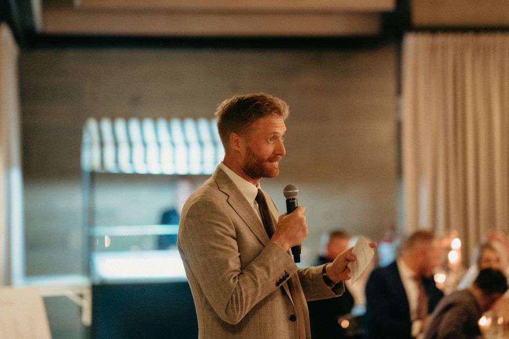 A speaker in a beige suit addresses the wedding guests during a reception.