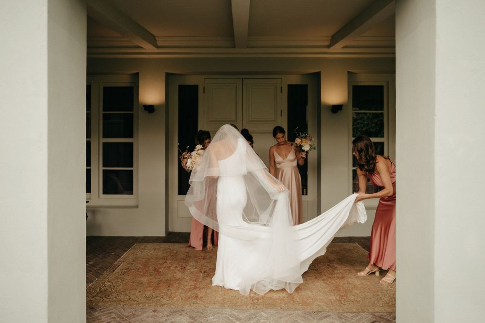 A bride in a white wedding dress with flowing veil being helped by attendants in a covered walkway.