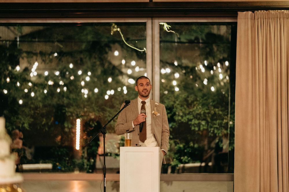 A speaker at a wedding reception stands at podium with twinkling lights in background.