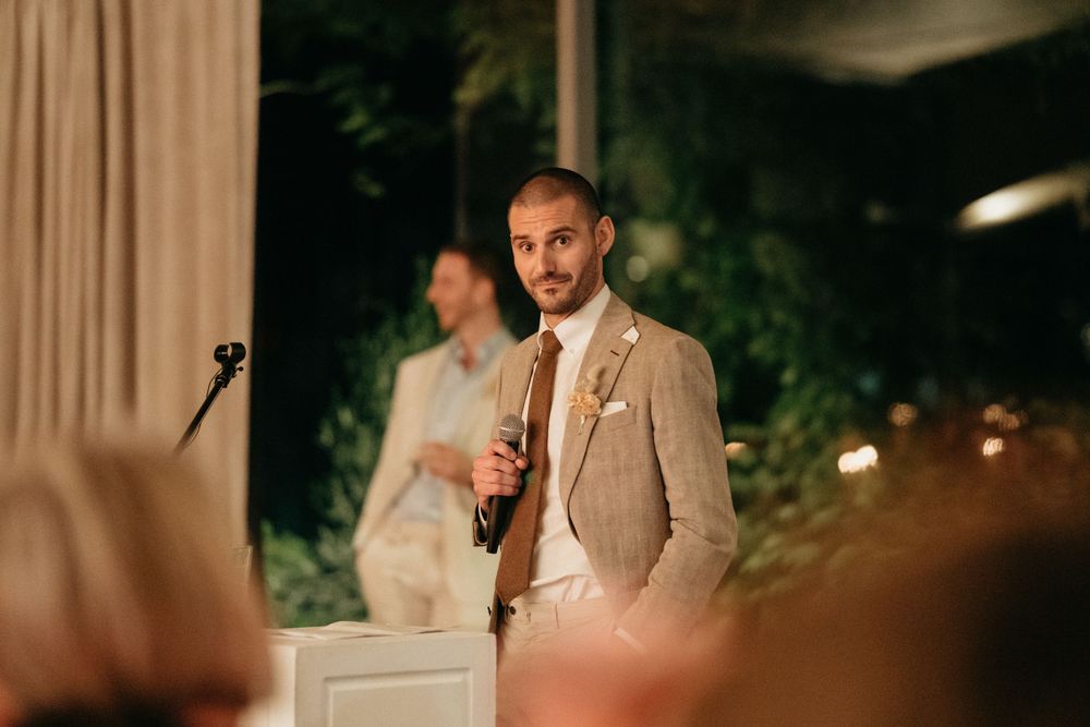 A person in a tan suit gives a wedding speech in front of foliage and curtains at an evening reception.