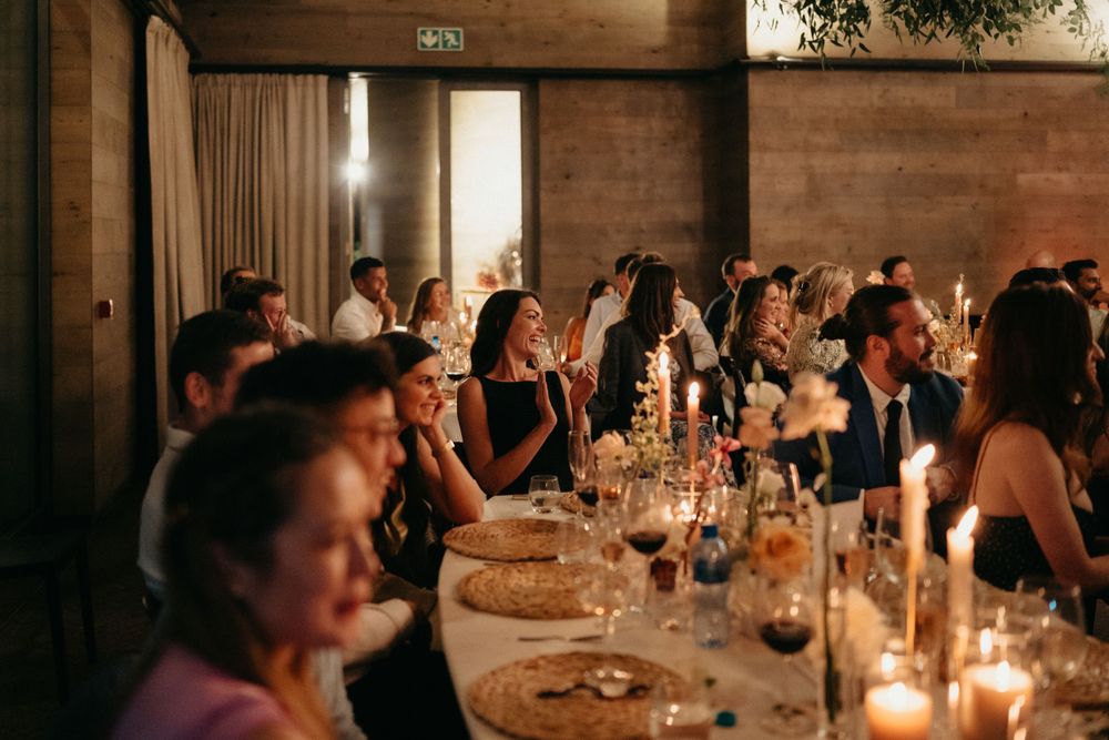 Wedding reception guests seated at long tables with glowing candles in a rustic venue.