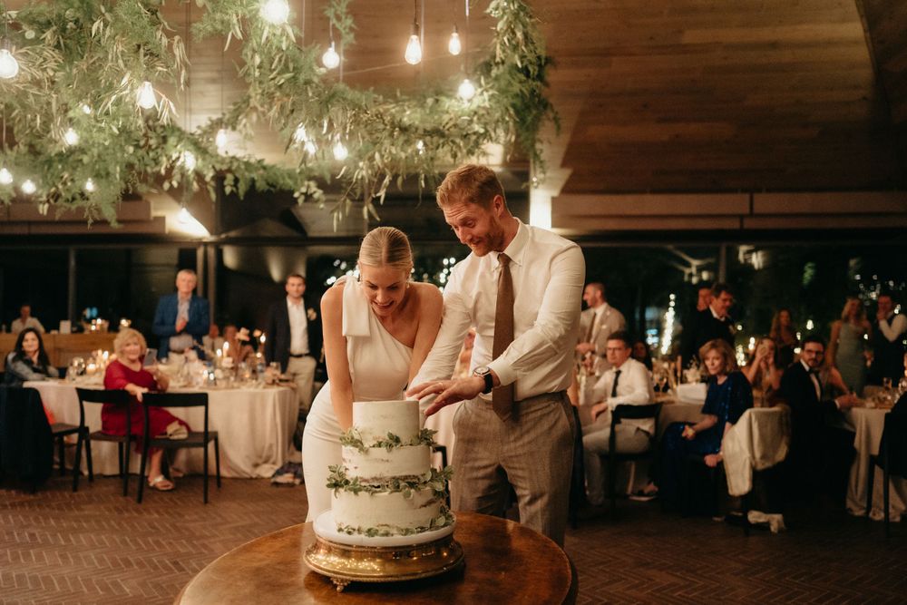 A couple cuts their wedding cake together at a rustic reception venue decorated with twinkling lights and greenery.