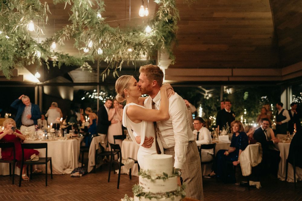 Newlyweds share their first dance surrounded by guests and hanging greenery at reception.