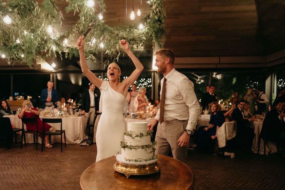 Newlyweds joyfully cutting their elegant tiered wedding cake under romantic string lights.