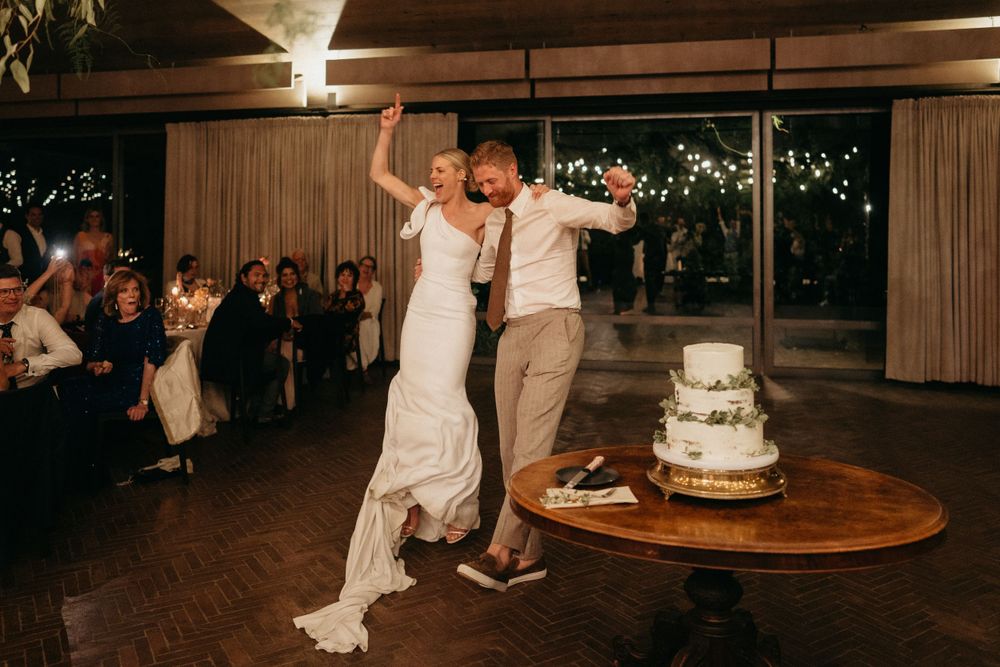 Newlyweds celebrate with joyful dance moves next to their wedding cake at an indoor reception venue.