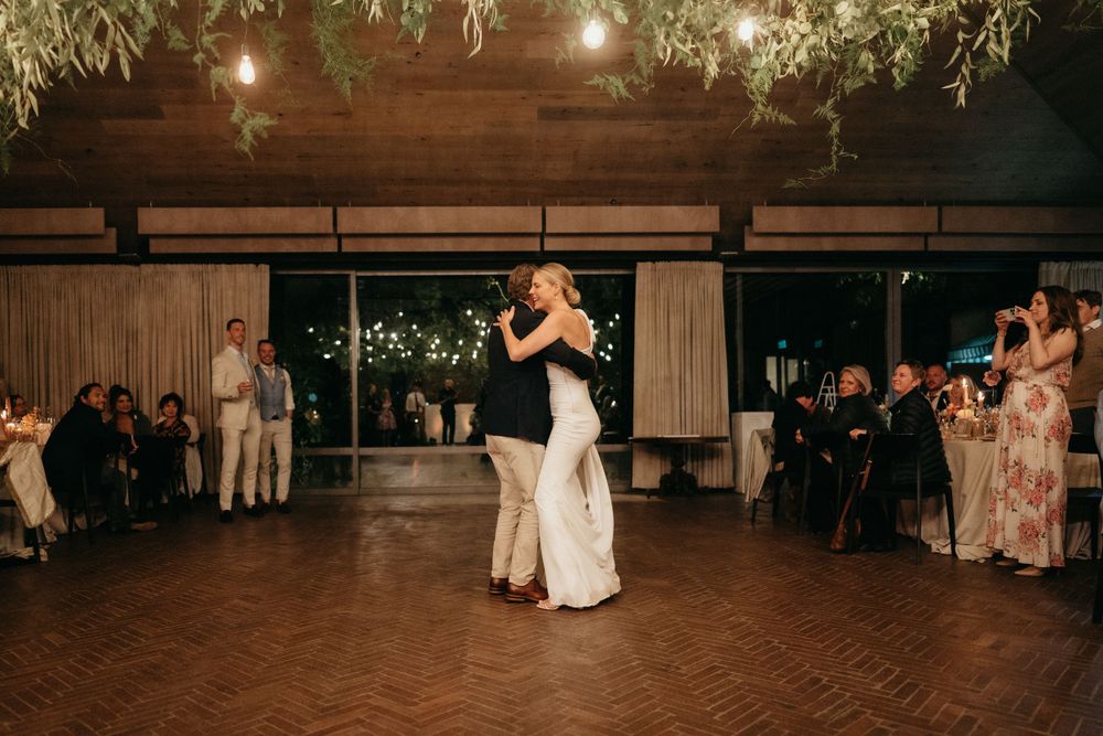 Newlyweds dance together in a rustic wooden venue while guests watch from the sides.