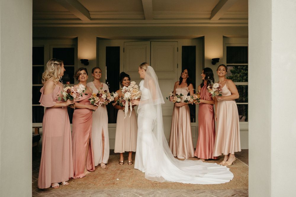 Bridesmaids in pink dresses gather around bride in white gown on covered porch.