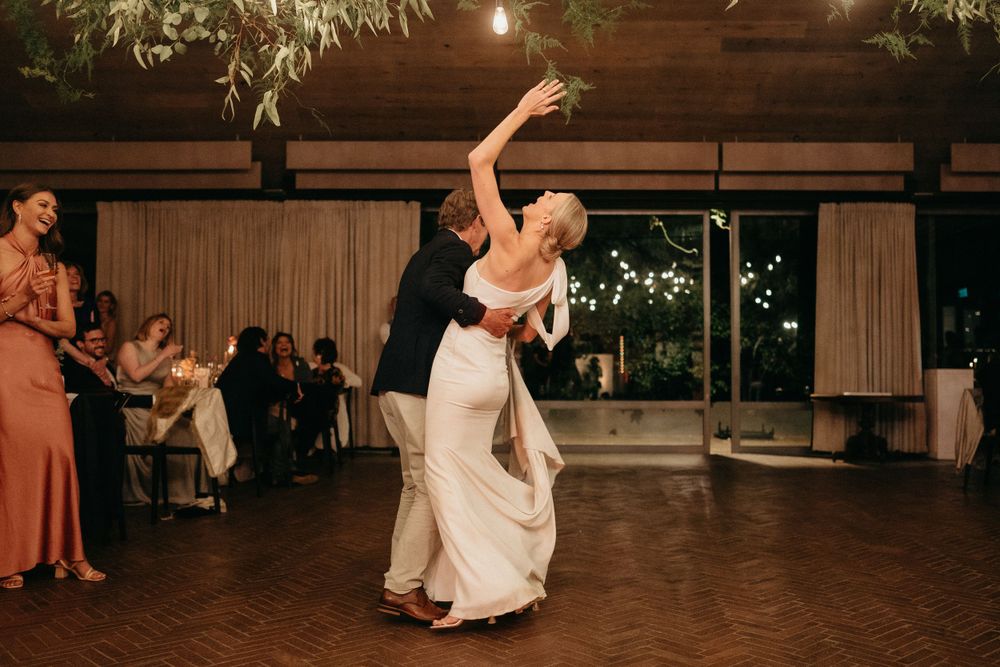 A couple performs an elegant dance move during their first dance at a wedding reception with guests watching.
