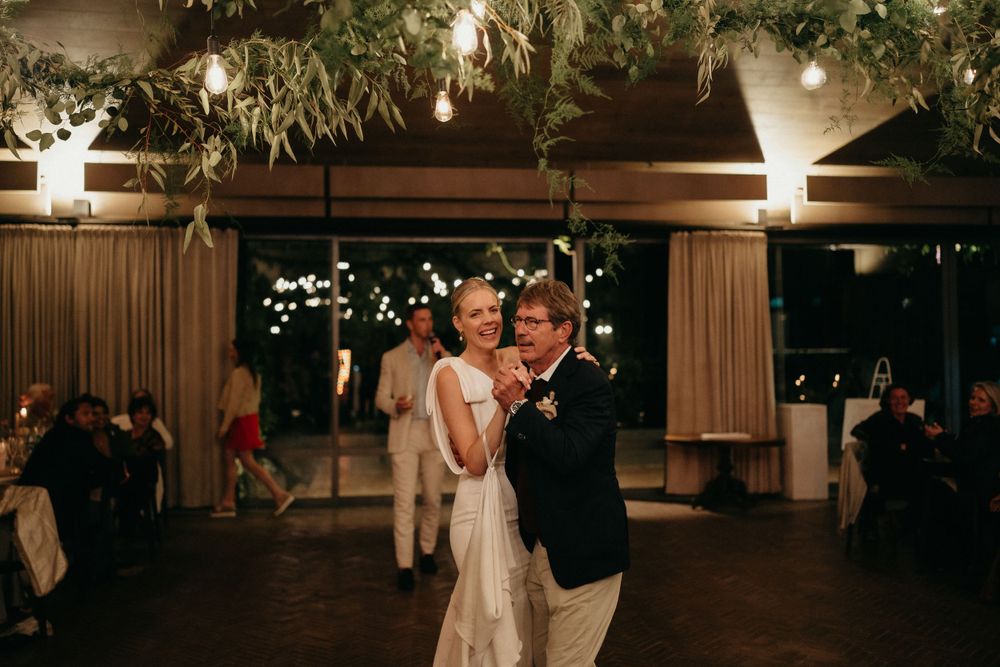 A couple shares their first dance at a wedding reception under twinkling lights and greenery.