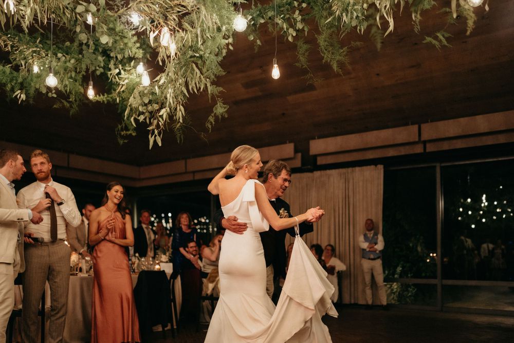 Wedding celebration in a rustic venue with greenery hanging from wooden beams and twinkling lights.