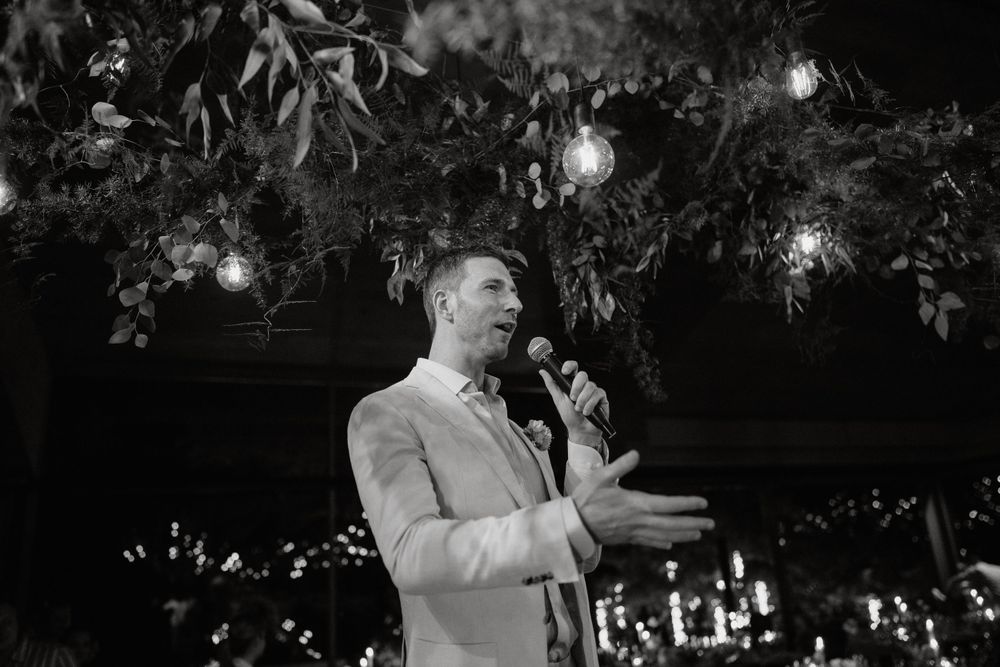 Black and white photo of speaker at wedding reception with hanging lights and greenery decor.