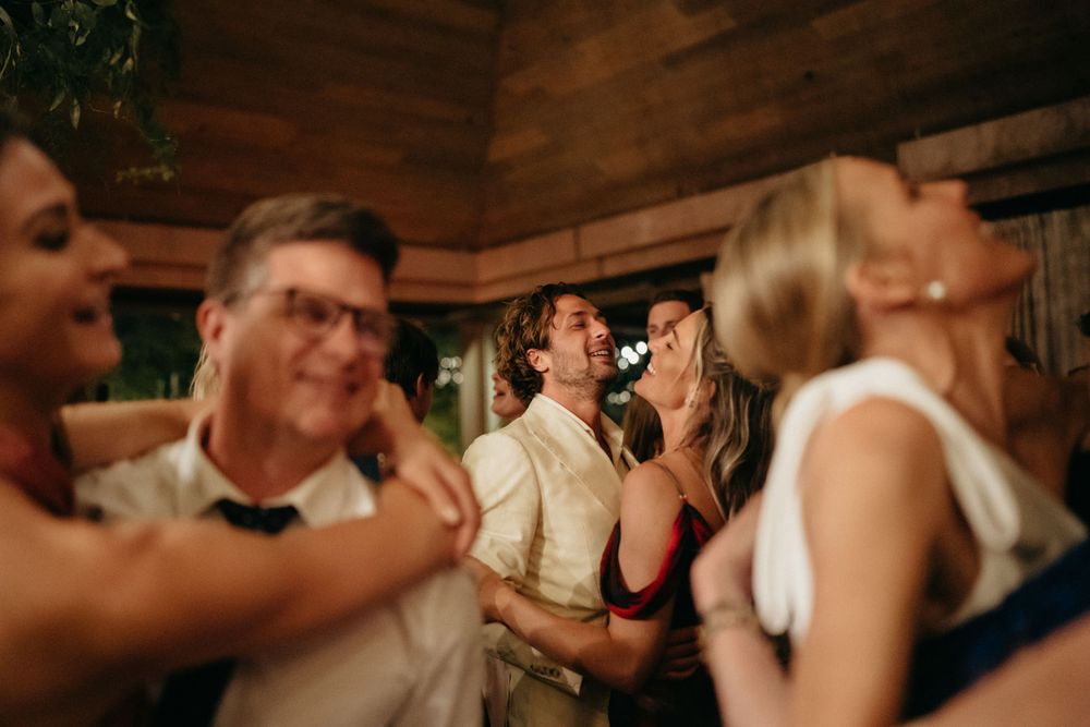 Wedding guests dance and celebrate on a crowded dance floor inside a rustic wooden venue.
