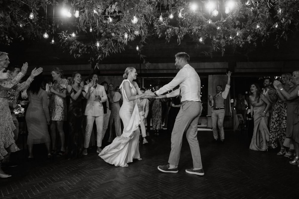 Newlyweds dance energetically on a dark dance floor under hanging lights while guests cheer.