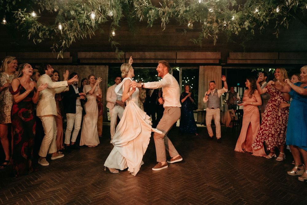 Newlyweds perform their first dance surrounded by guests on a wooden dance floor.