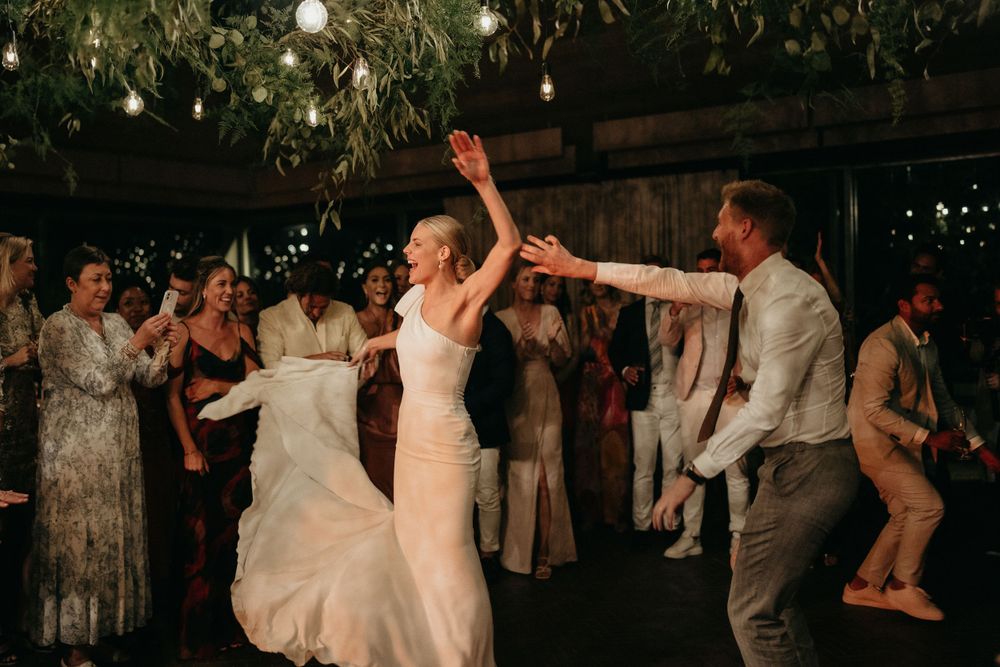 Couple dancing at wedding reception under string lights with guests watching and celebrating.